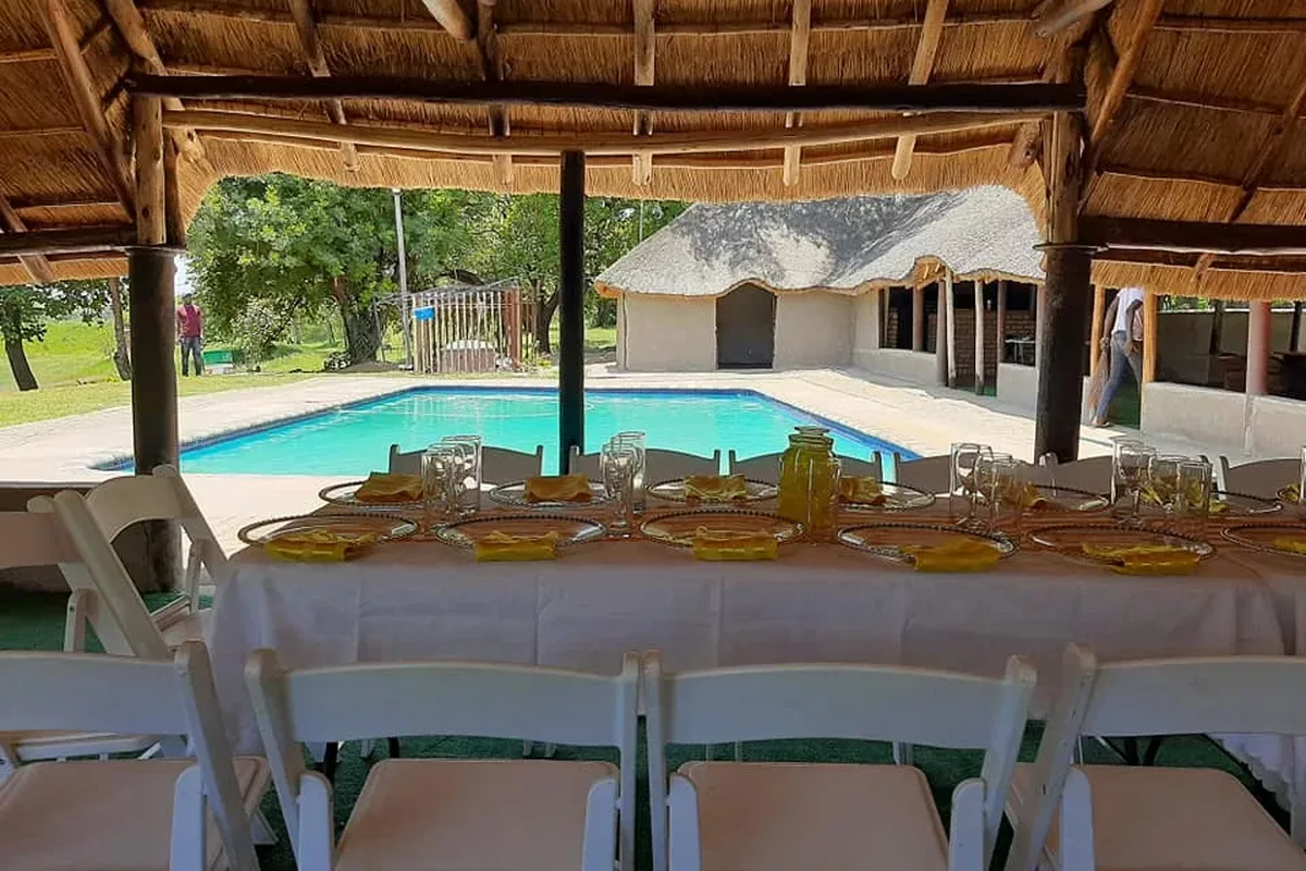 Dining table setup under a thatched roof next to a swimming pool at Refilwe Farm Resort in Tzaneen